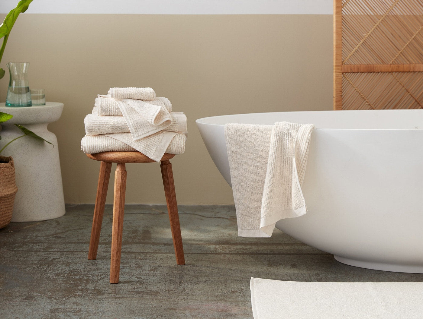 Coyuchi Temescal Ribbed towels stacked on wooden stool beside freestanding bathtub in ivory.