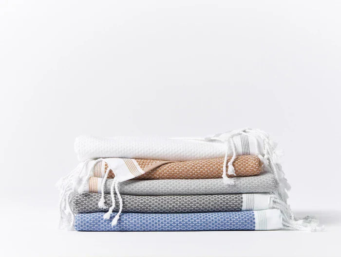 Stack of Coyuchi Mediterranean guest towels in white, camel, grey, and blue with fringe trim.