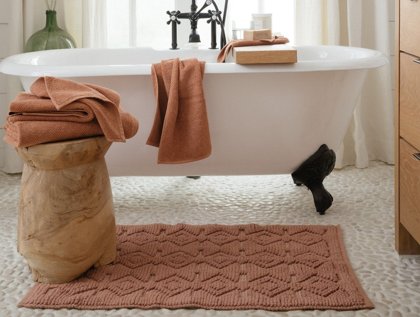Coyuchi Mosaic Canyon bath rug in clay, textured diamond pattern, styled on pebble floor beside clawfoot tub.