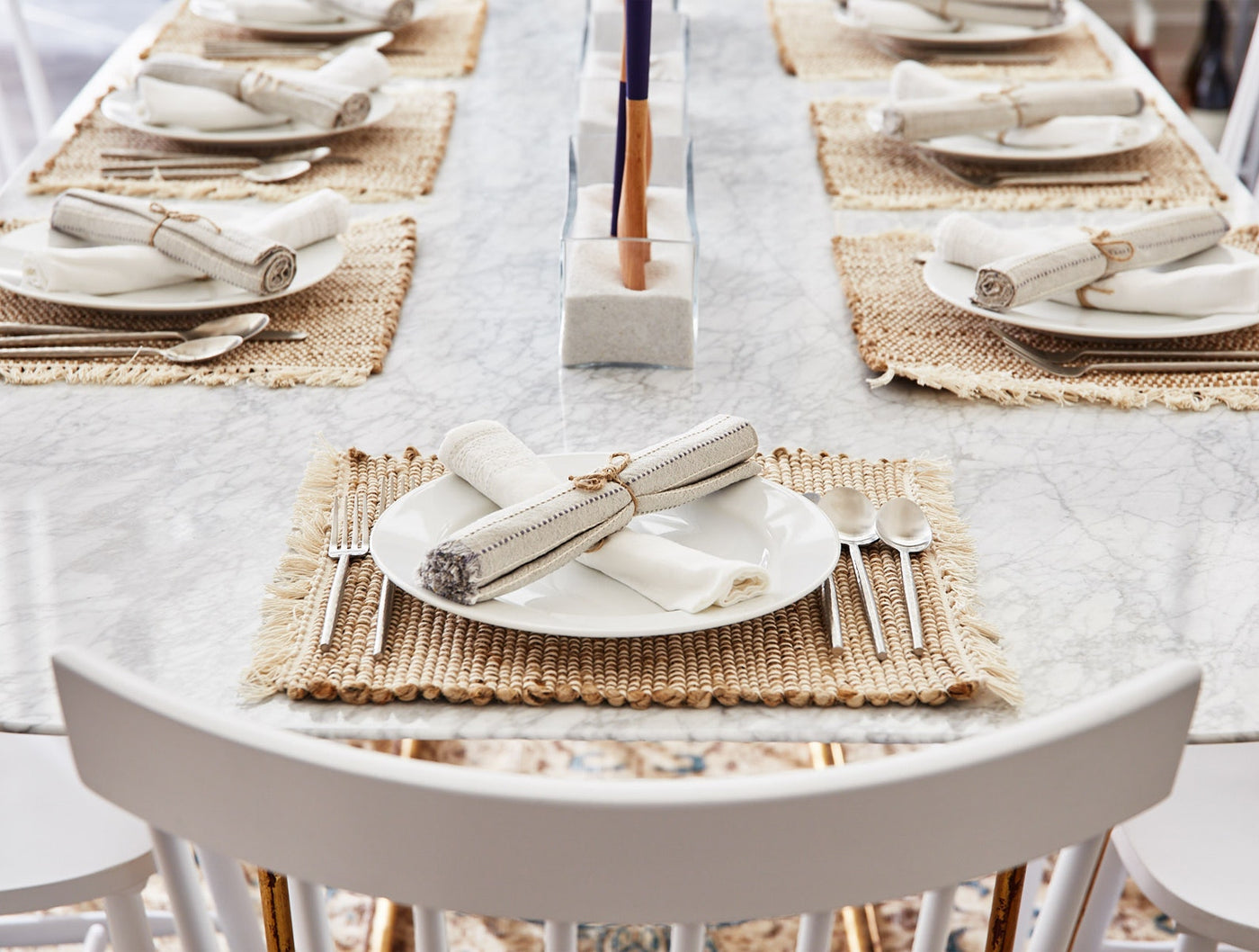 Rolled ivory napkins tied with twine on white plates atop woven jute placemats, marble table setting.