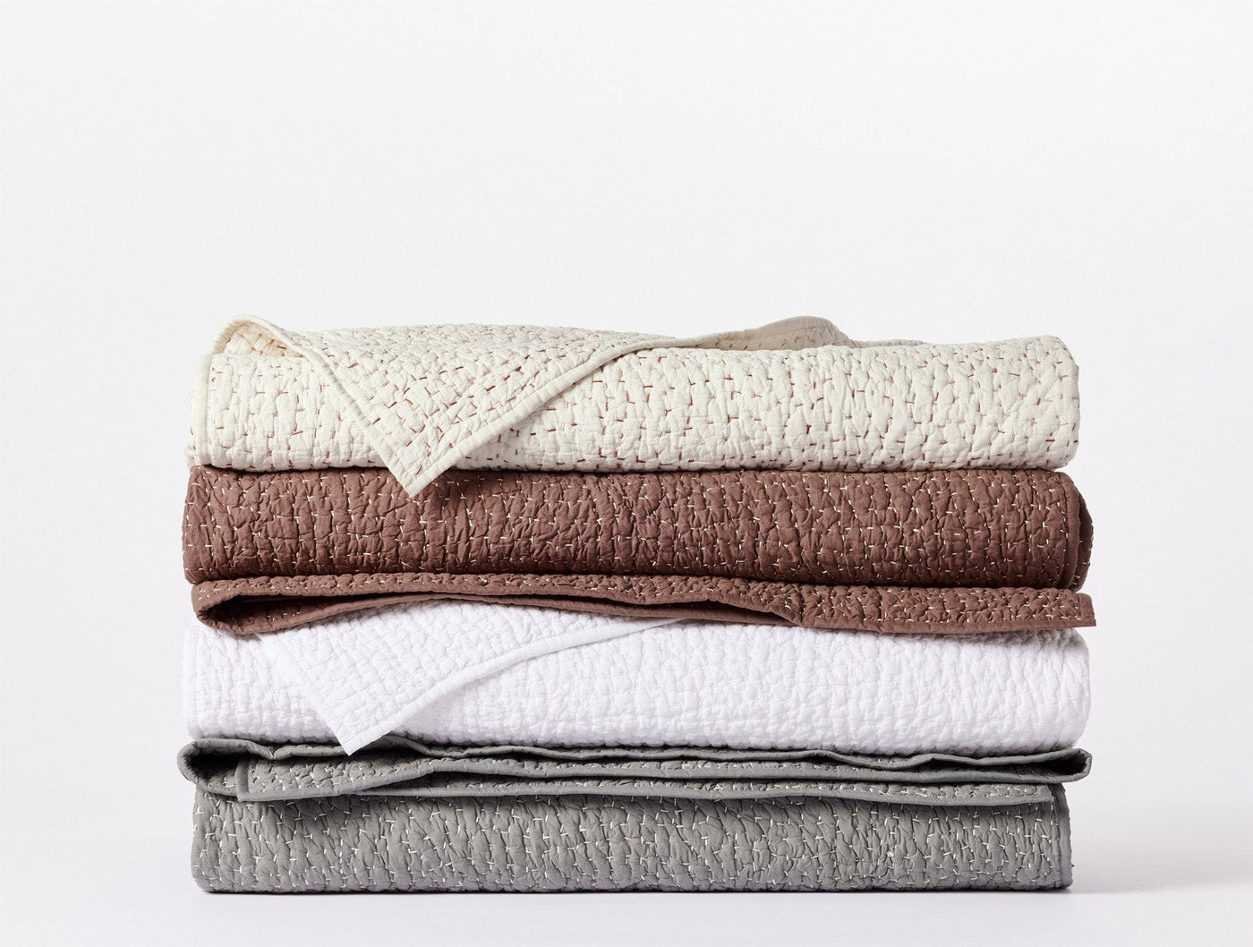 Stack of folded Coyuchi Pebbled Handstitched quilts in ivory, brown, white, and grey colorways.