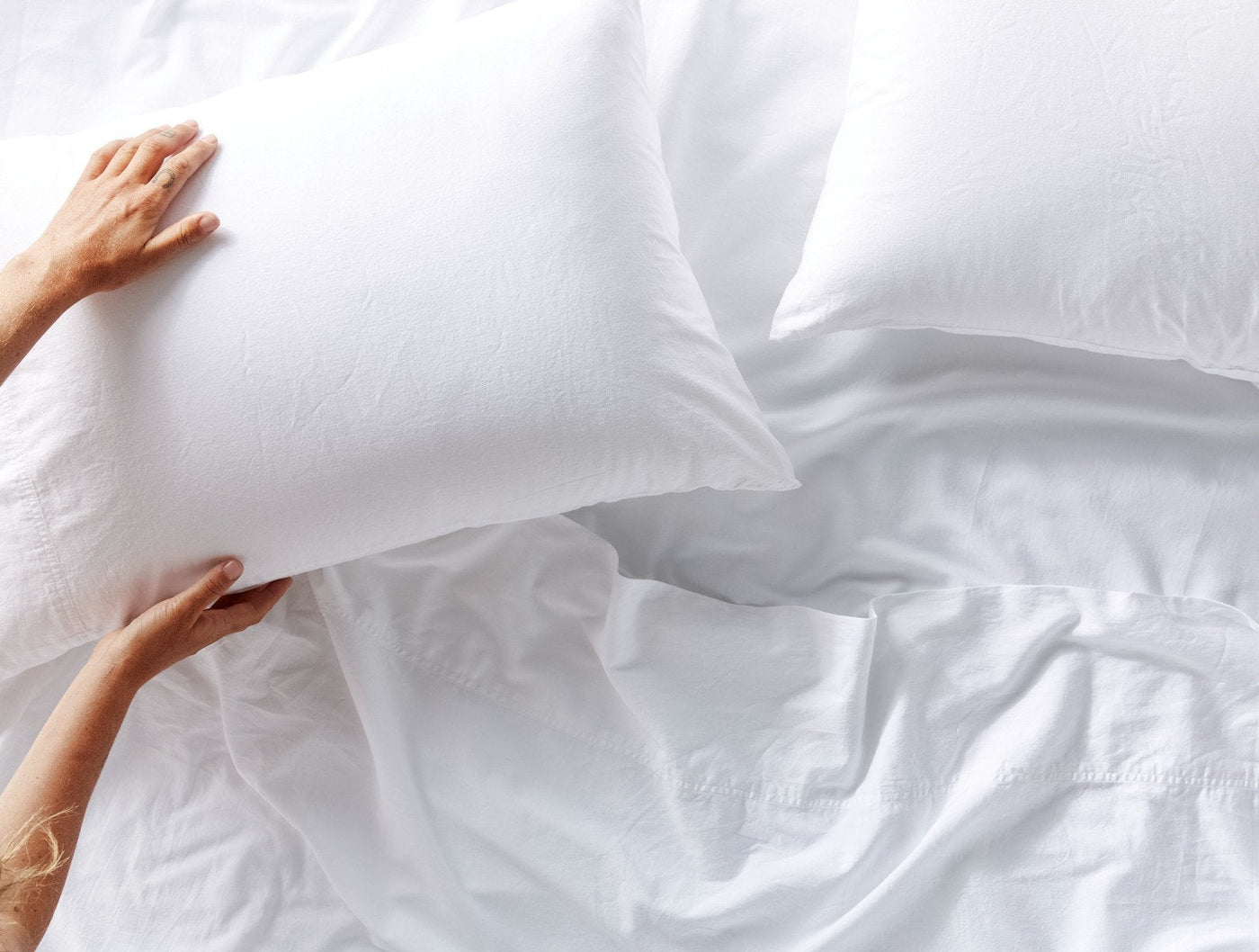 Hands arranging a white Coyuchi Cloud Soft Sateen pillowcase on a softly rumpled white bed.