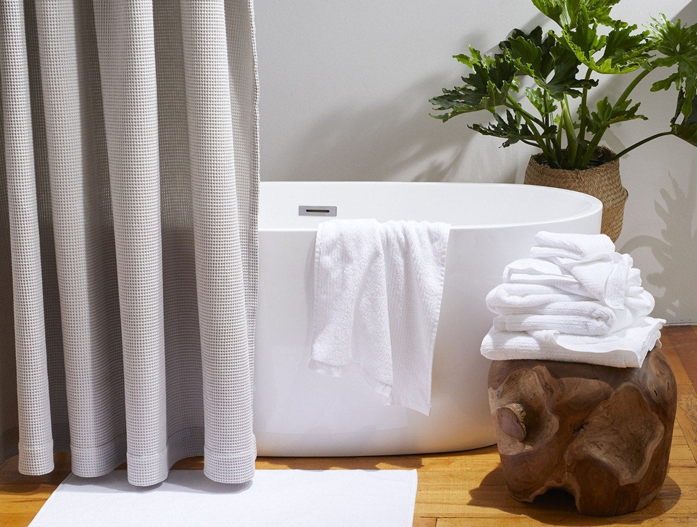 White ribbed bath towel draped over freestanding tub, stacked towels on wood stump beside green plant.
