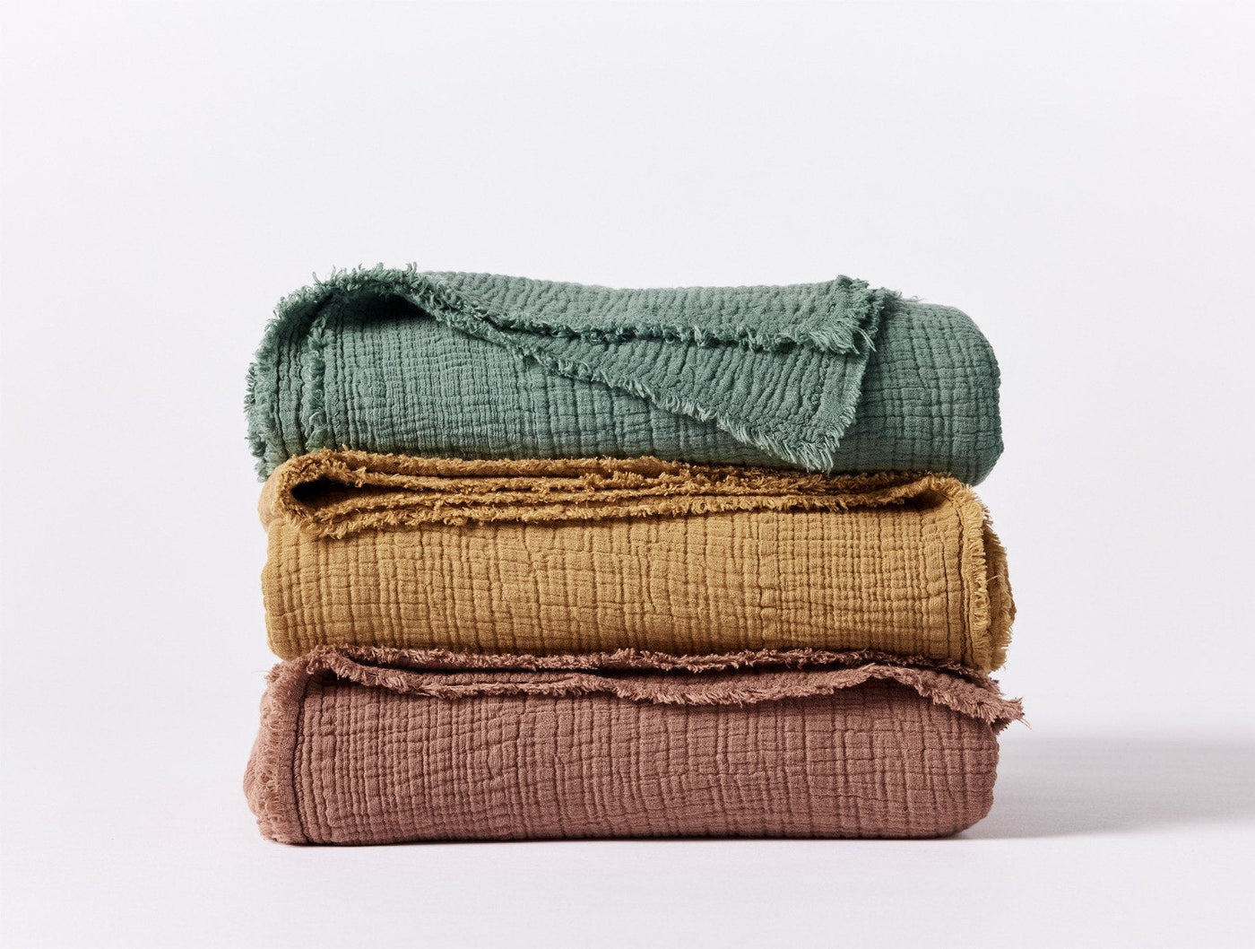 Three stacked Coyuchi Topanga Matelasse throws in sage, ochre, and dusty rose with fringe trim.
