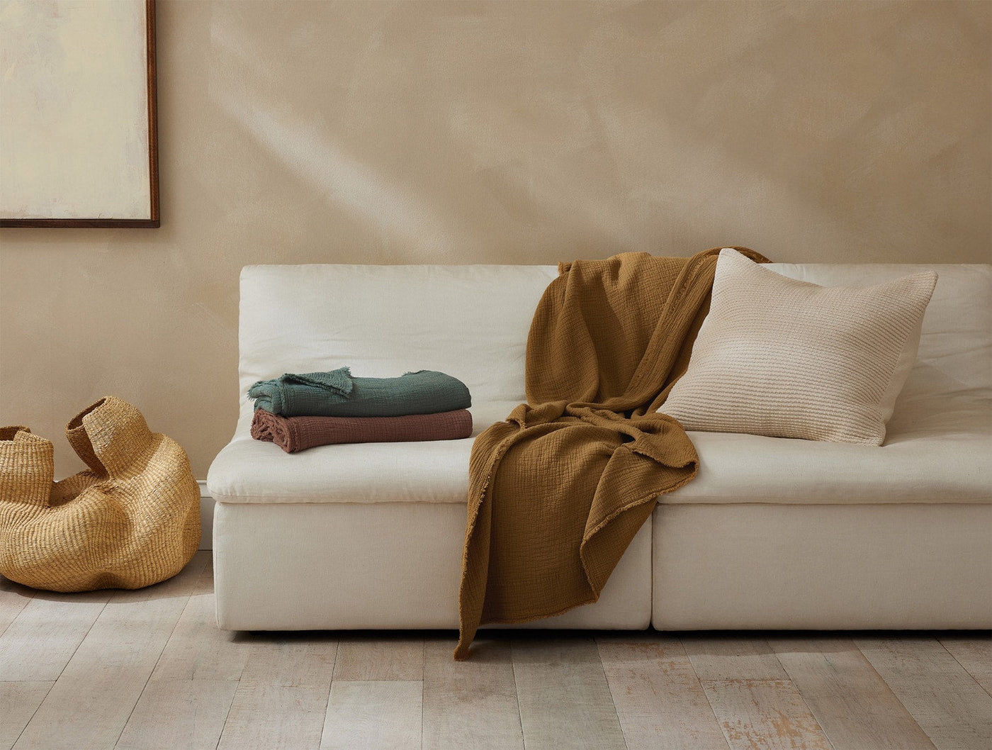 Ochre throw draped over a cream sofa beside folded sage and mauve throws in a warm neutral room.