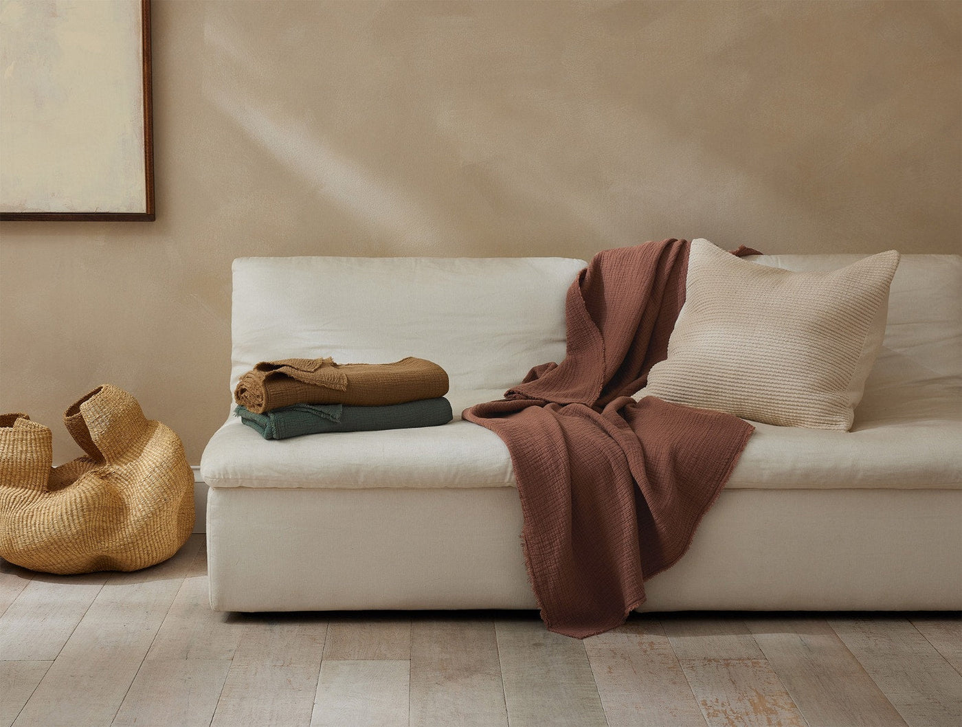 Rust-colored matelasse throw draped over a cream sofa beside folded throws in olive and camel.