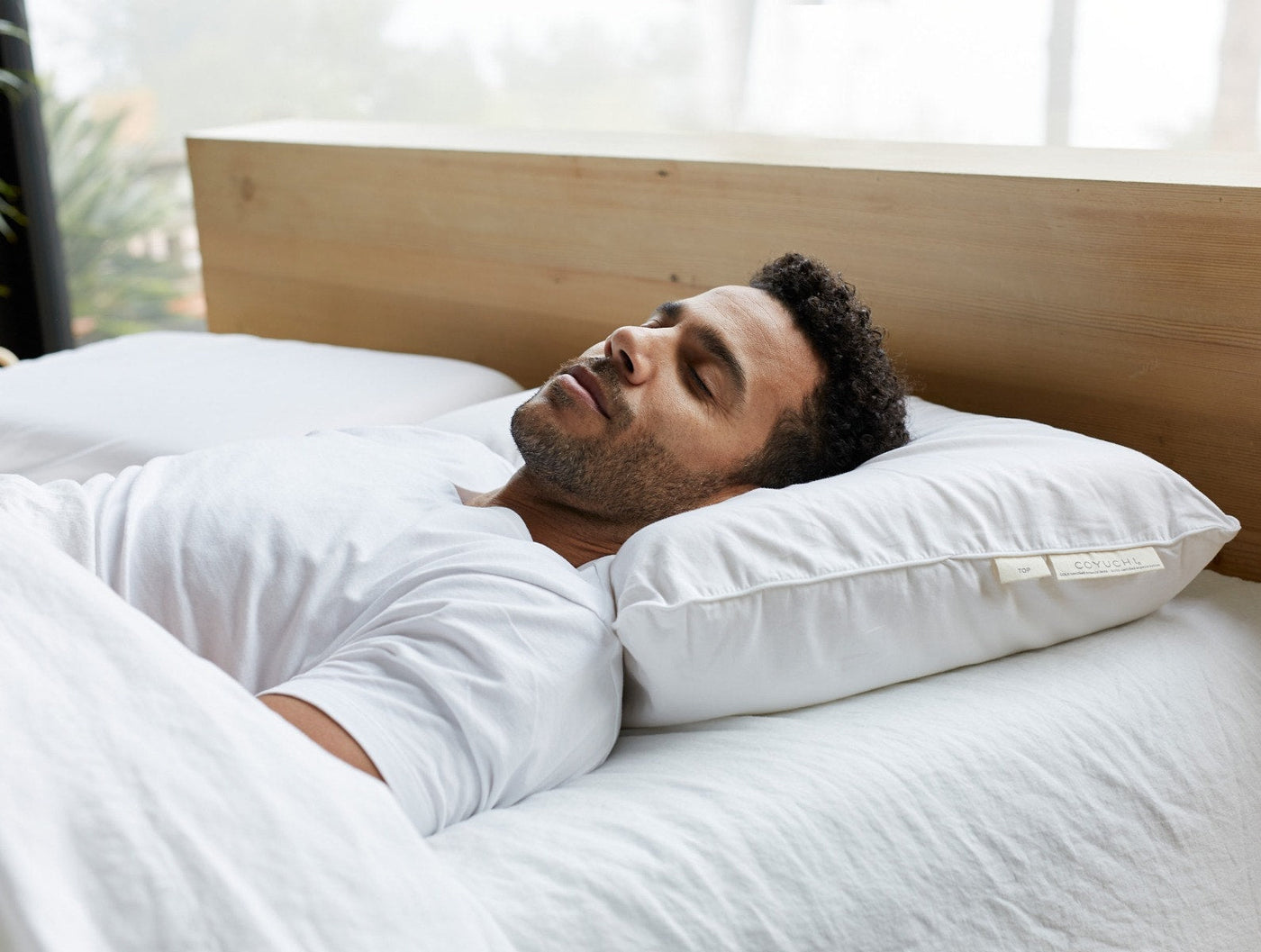 Man resting on a white Coyuchi Turiya Latex pillow in a bright bedroom setting.