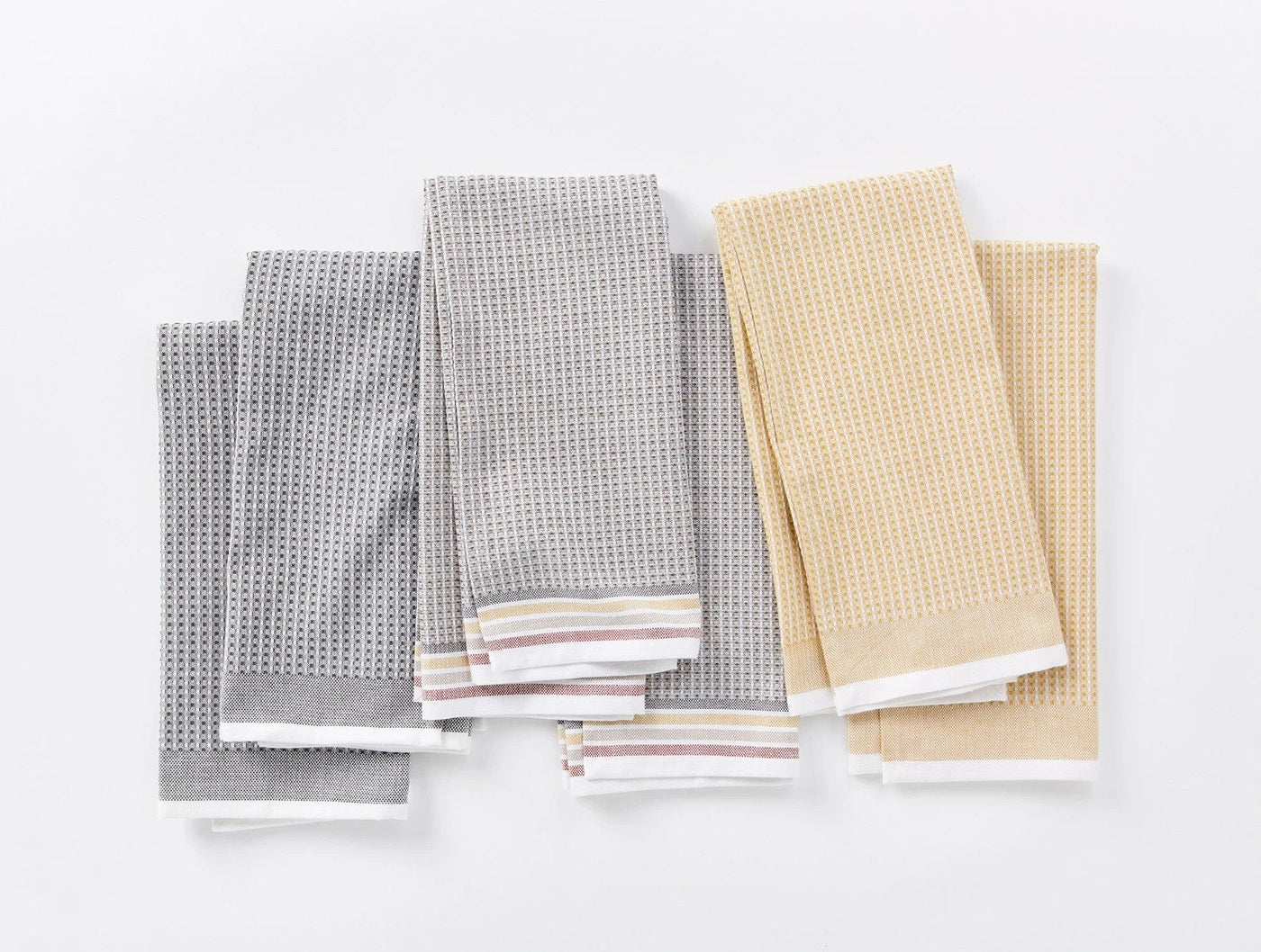 Six Coyuchi Waffle kitchen towels folded flat, three grey and three gold, with striped hems.