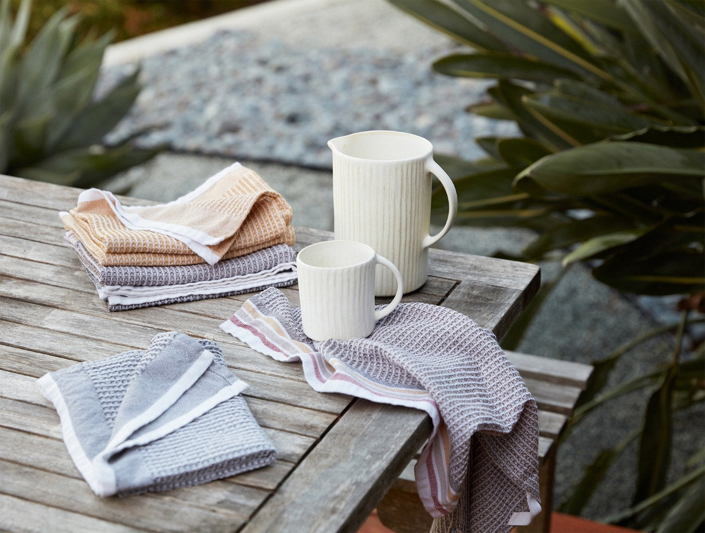 Coyuchi waffle kitchen towels in muted tones styled on a weathered wood table outdoors.