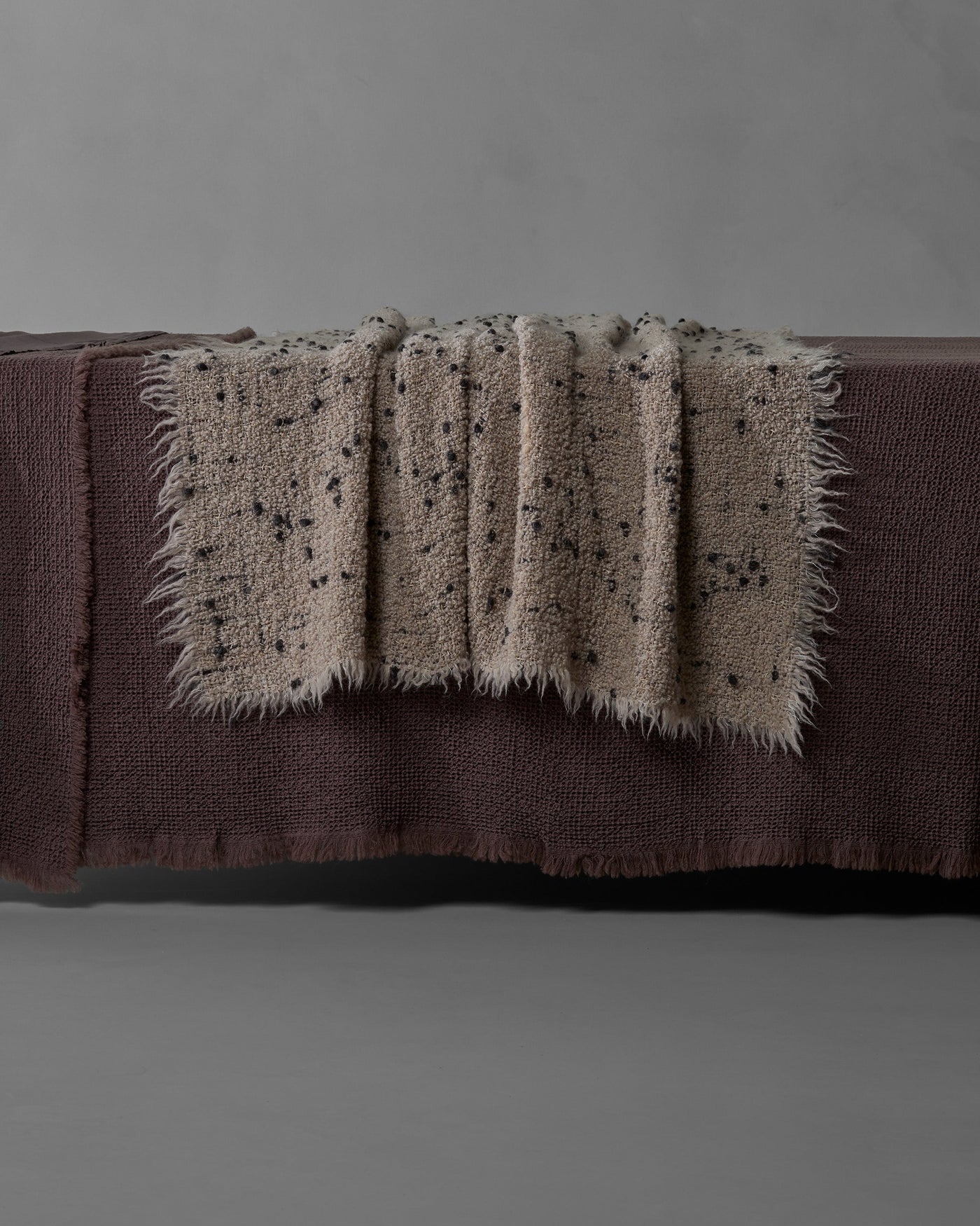 Fringe-edged peg throw in taupe with black speckled texture, draped over a plum linen bench.