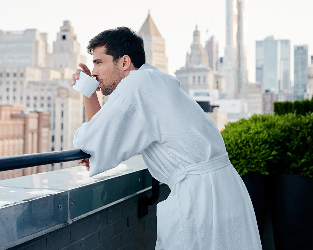 A man in Frette’s The Pique Kimono stands on a balcony, sipping from a white mug as city buildings rise in the background.