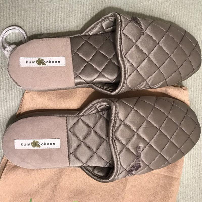A pair of gray Quilted Silk Slippers by Kumi Kookoon are displayed on beige fabric, offering a luxurious spa-like comfort at home.