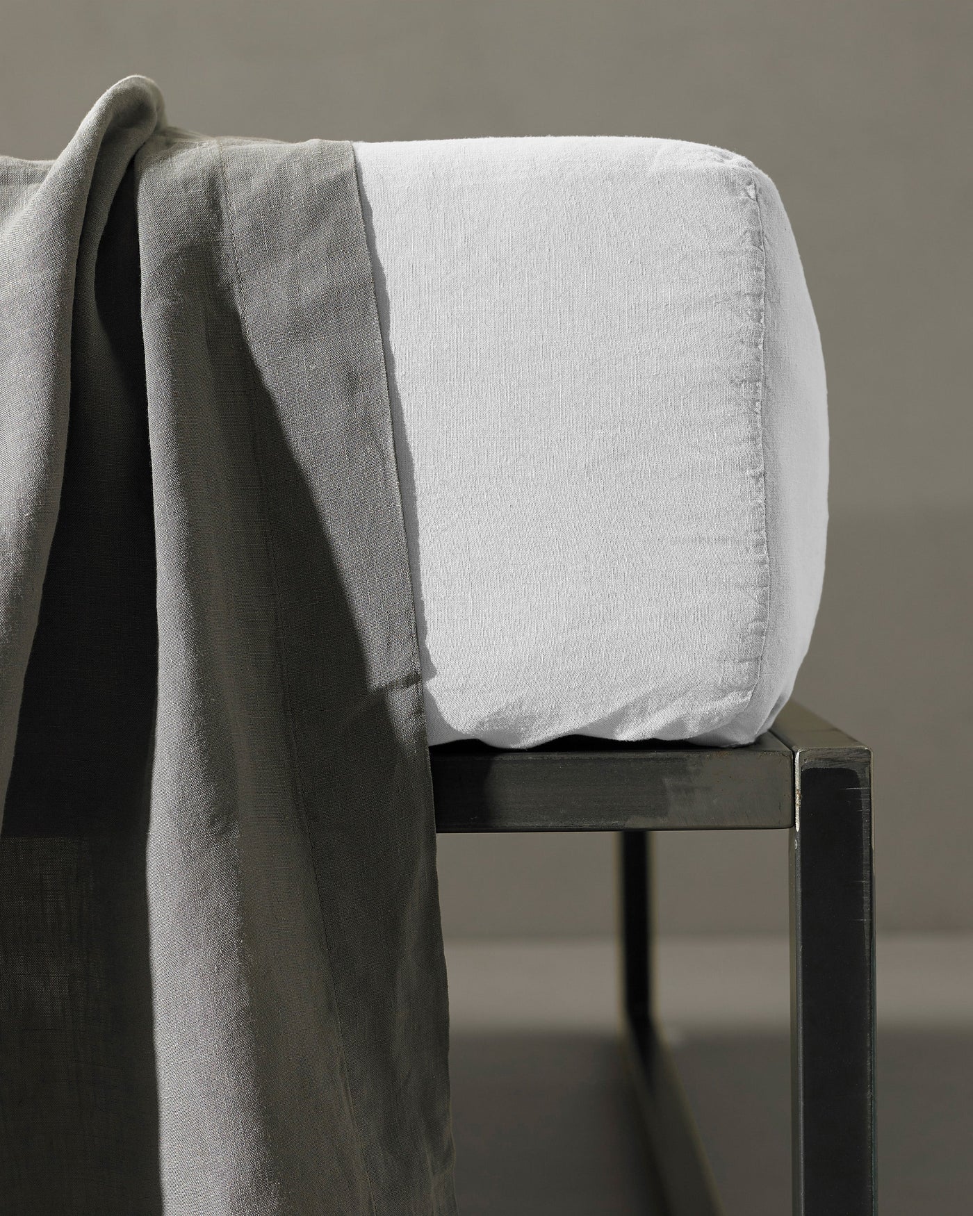 White fitted sheet corner detail draped over a dark chair with grey linen fabric.