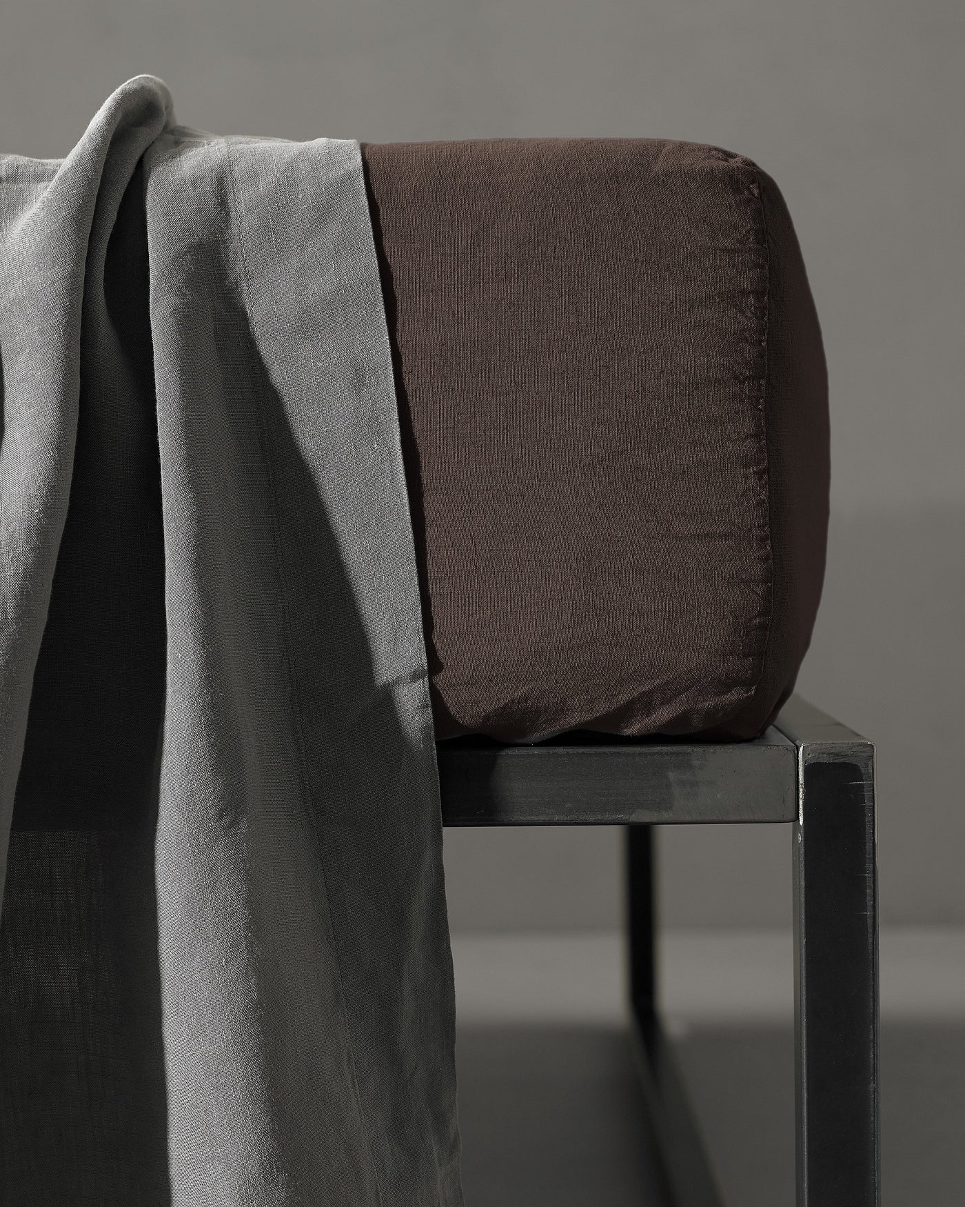 Espresso fitted sheet corner detail draped over dark metal bench with grey linen throw.