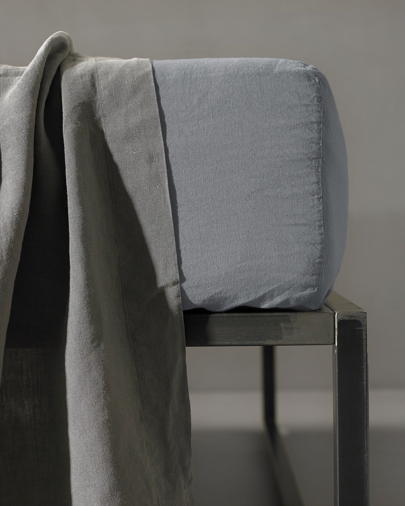 Slate fitted sheet corner detail draped over a dark metal stool with linen fabric alongside.