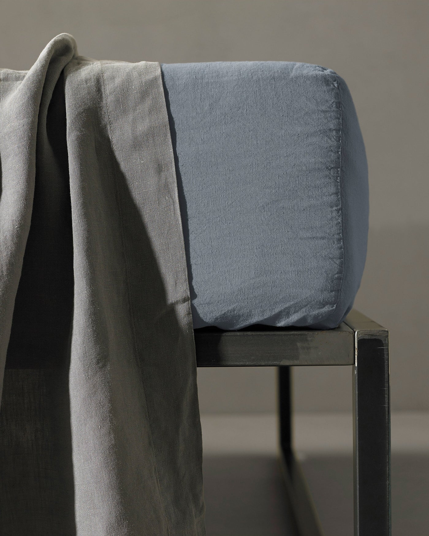 Society Limonta Rem fitted sheet in slate blue on a dark metal bench with grey linen draped alongside.