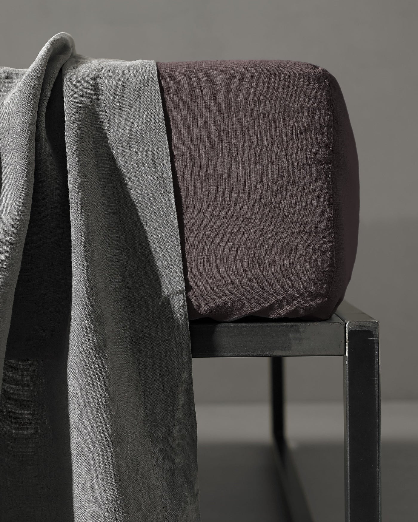 Charcoal fitted sheet on a bolster, styled with a draping ash linen cloth on a dark metal bench.