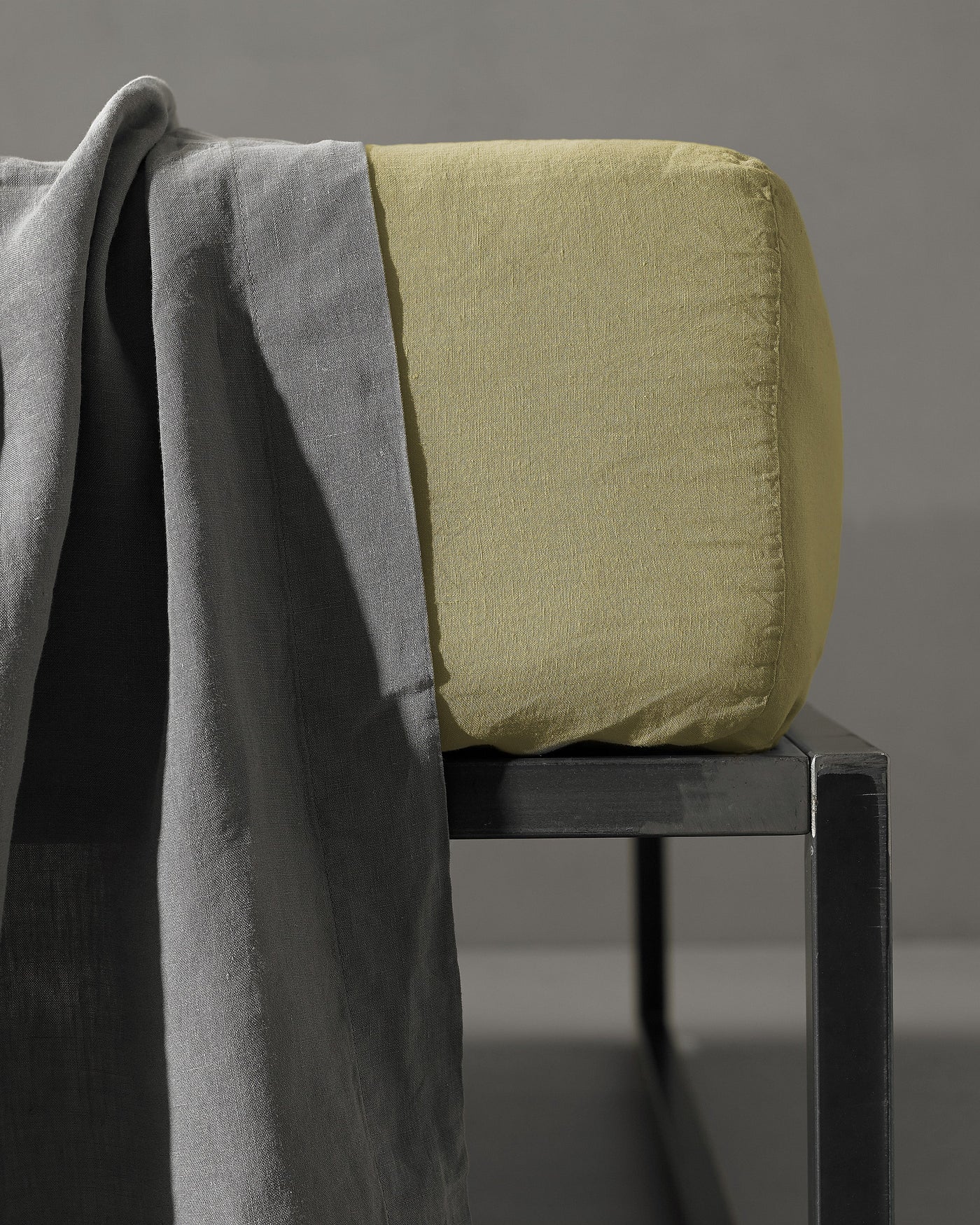 Sage fitted sheet corner detail on dark stool with slate linen draped alongside.