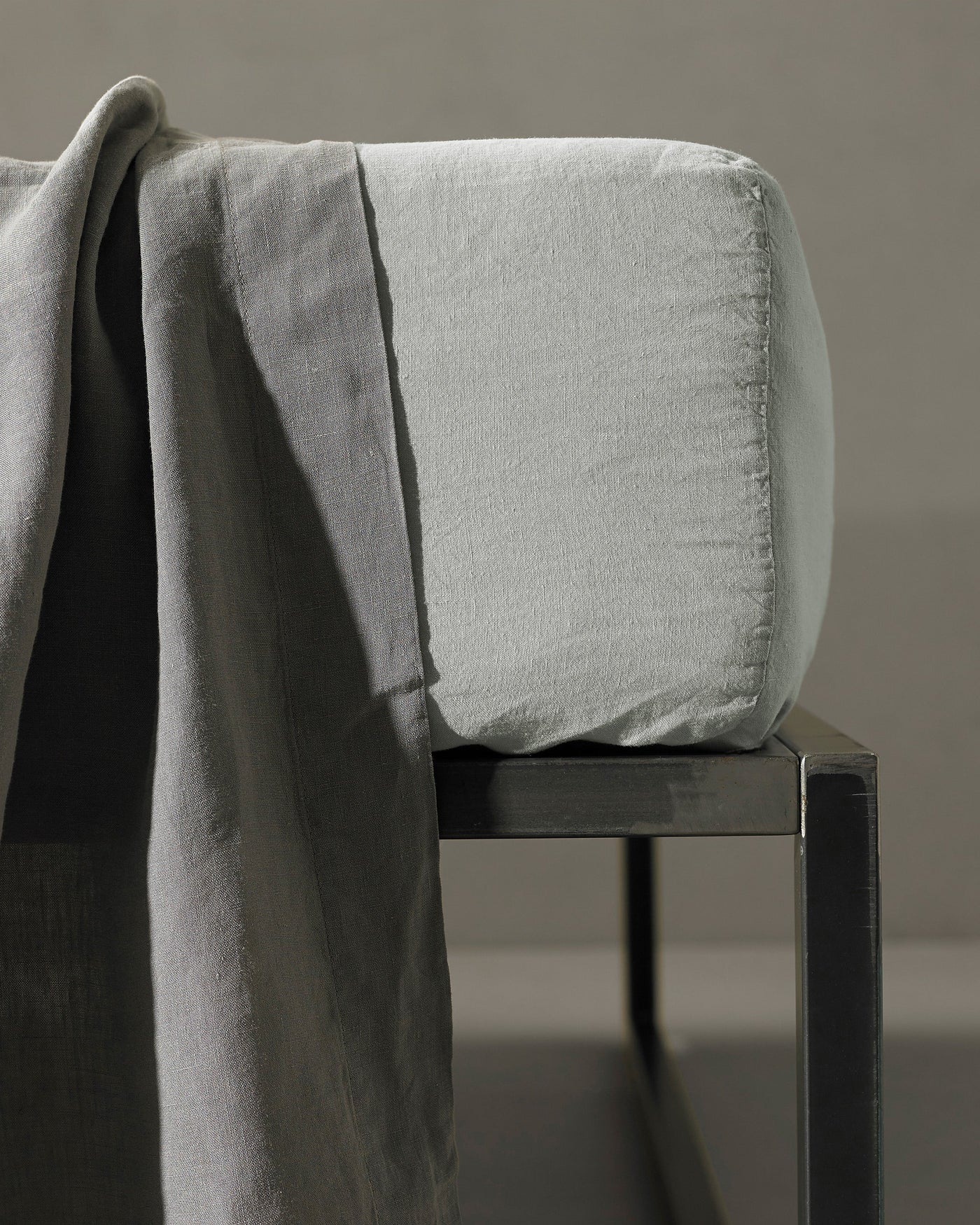 Dove grey fitted sheet corner detail showing relaxed linen texture on a dark chair.