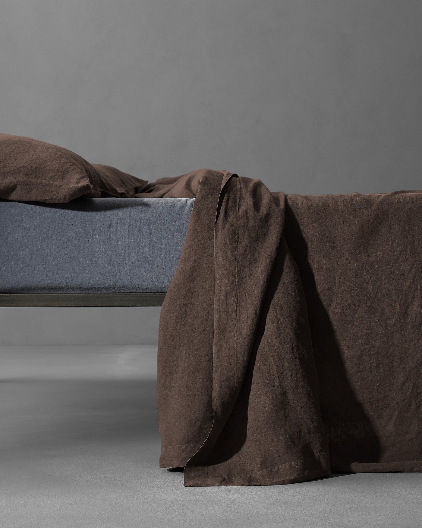 Society Limonta Rem flat sheet in espresso linen draped over bed edge against grey background.