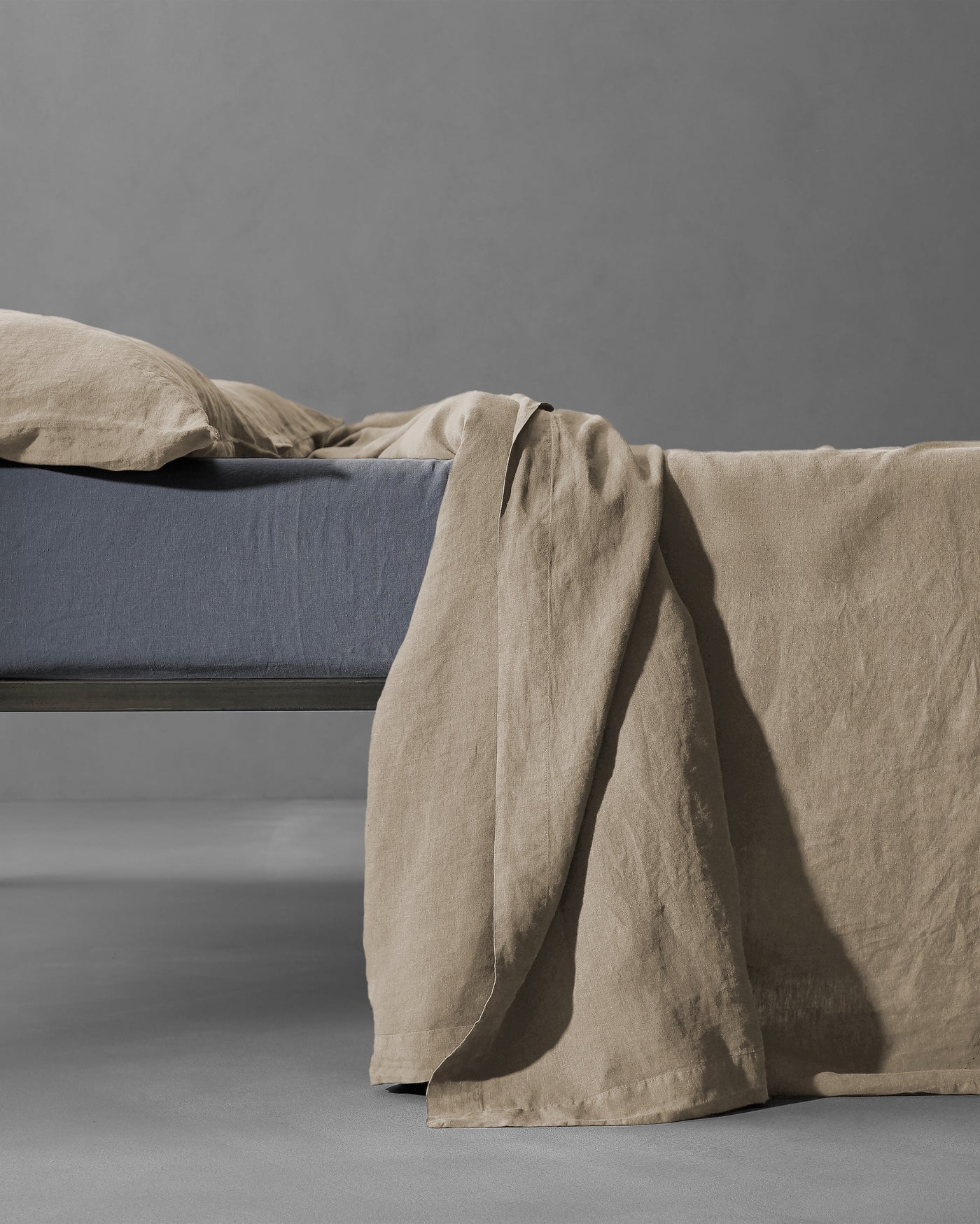 Sand linen flat sheet draped over bed edge, showing relaxed texture against slate fitted sheet.