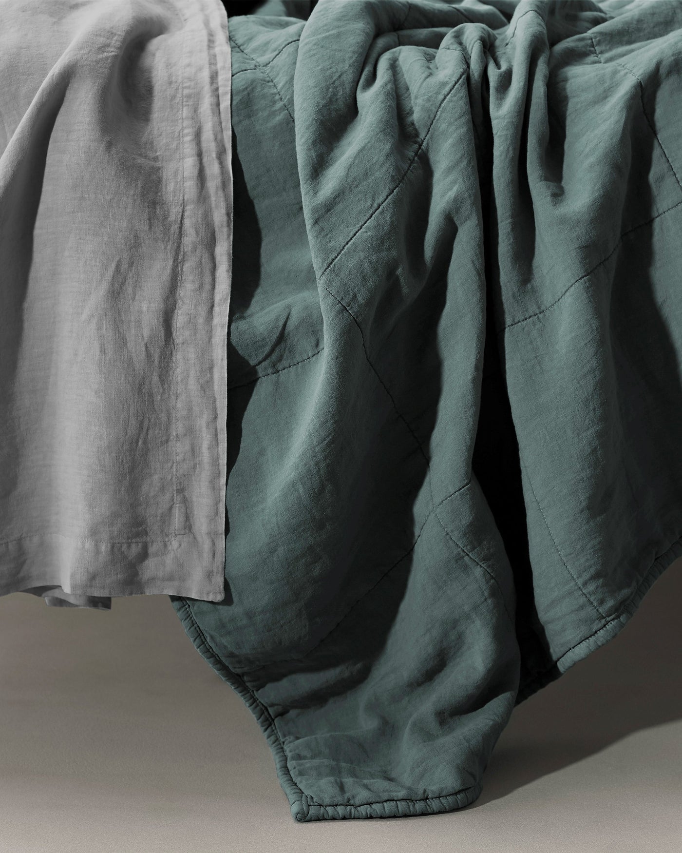 Teal linen quilt draped over bed edge alongside a grey linen sheet, showing quilted seam detail.