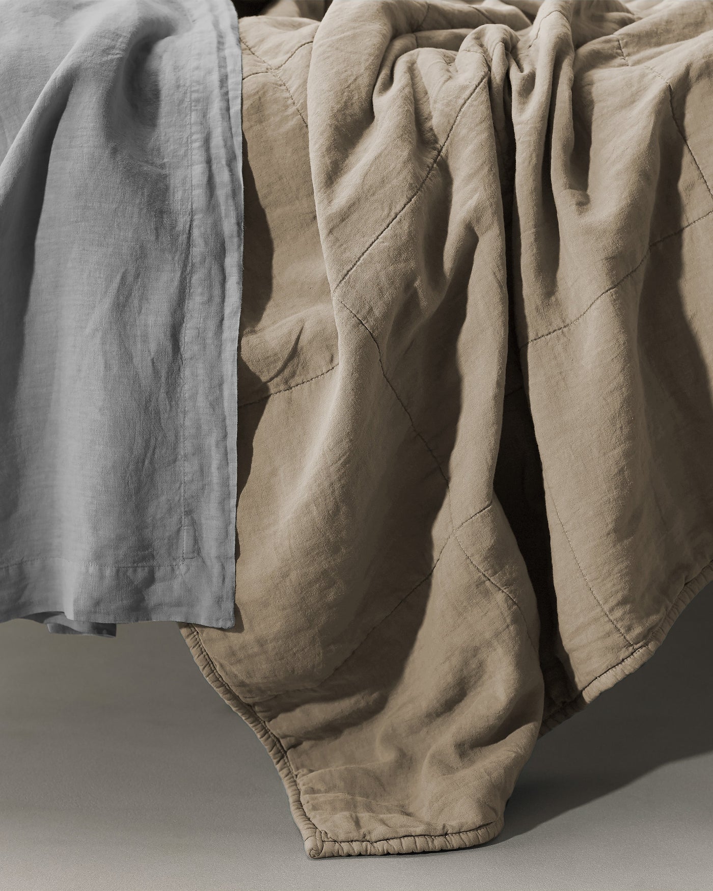 Society Limonta Rem quilt in sand linen draped over bed edge, diamond quilting detail visible.