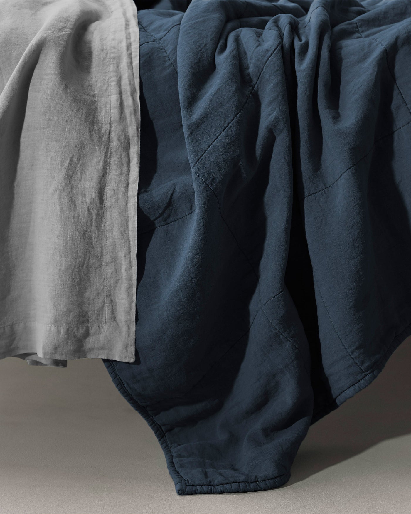 Society Limonta Rem quilt in navy linen draped over bed edge with silver-grey flat sheet.