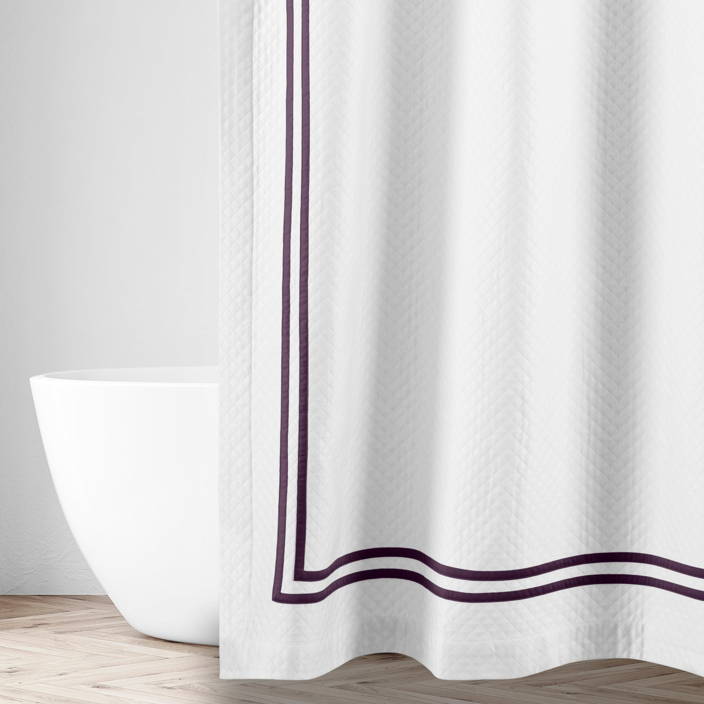 White matelassé shower curtain with double aubergine satin border trim, styled in a bright bathroom.