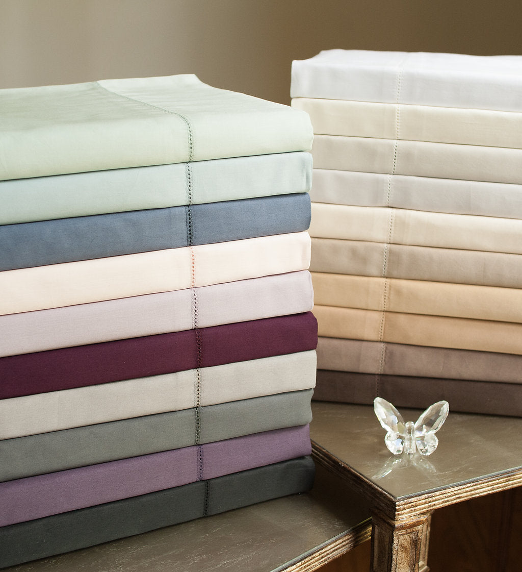 Two stacks of folded Home Treasures Royal sheets in multiple colorways with hemstitch detail.