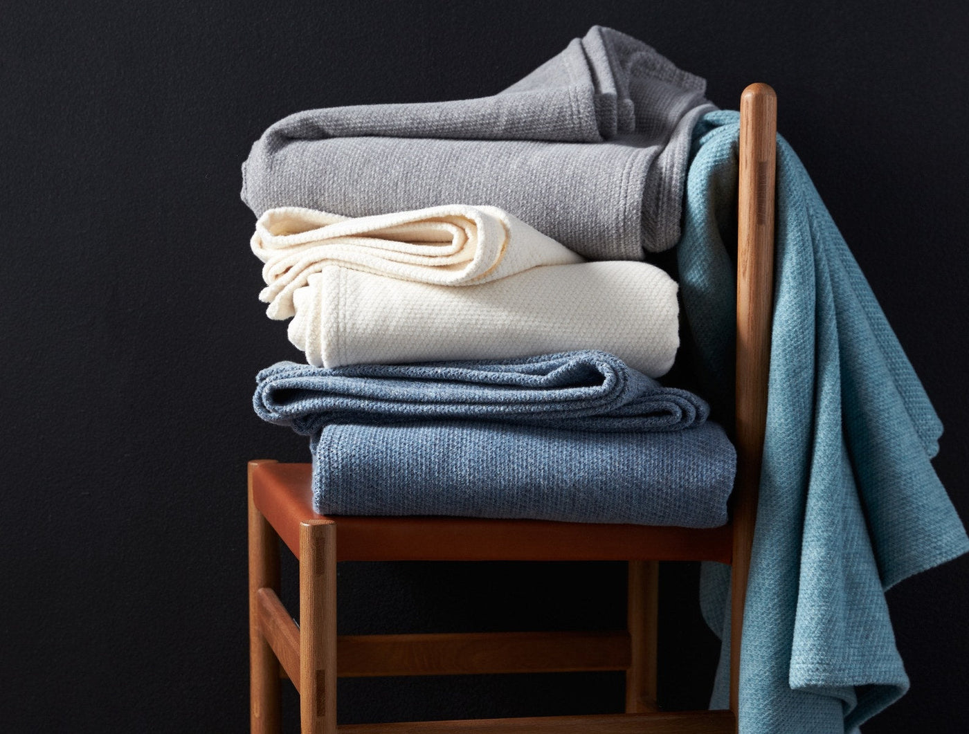 Stack of Coyuchi Sequoia throws in grey, ivory, slate blue, and teal draped on a wood chair.