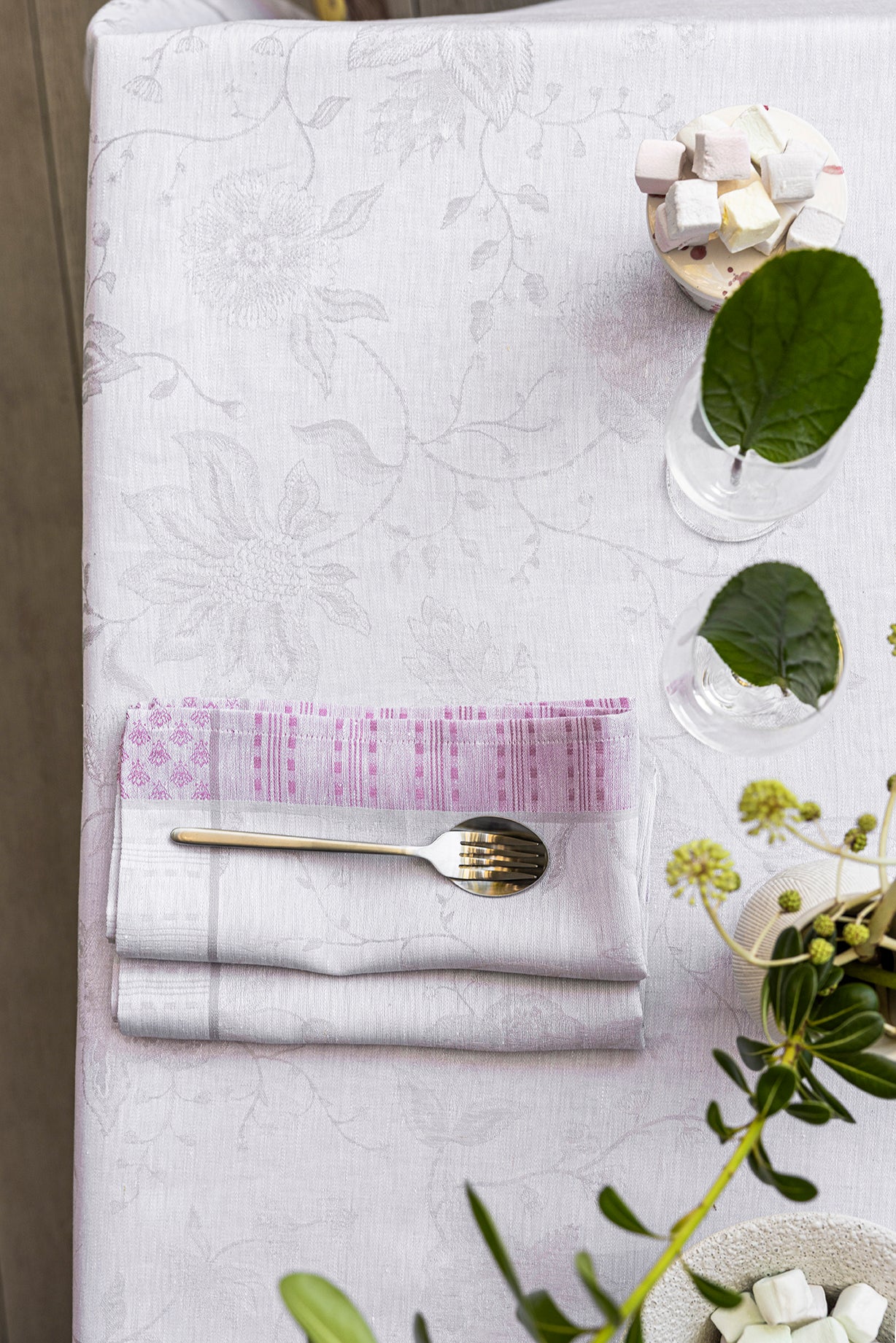 Close-up of folded white jacquard napkin with pink striped trim and gold fork on floral tablecloth.