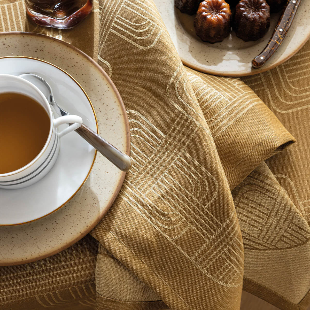 Le Jacquard Français Brown dinner napkin draped beside a teacup, showing woven geometric arc pattern.