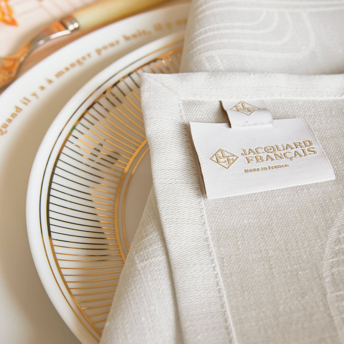 Gray Le Jacquard Français dinner napkin folded beside a gold-rimmed plate, brand label visible.