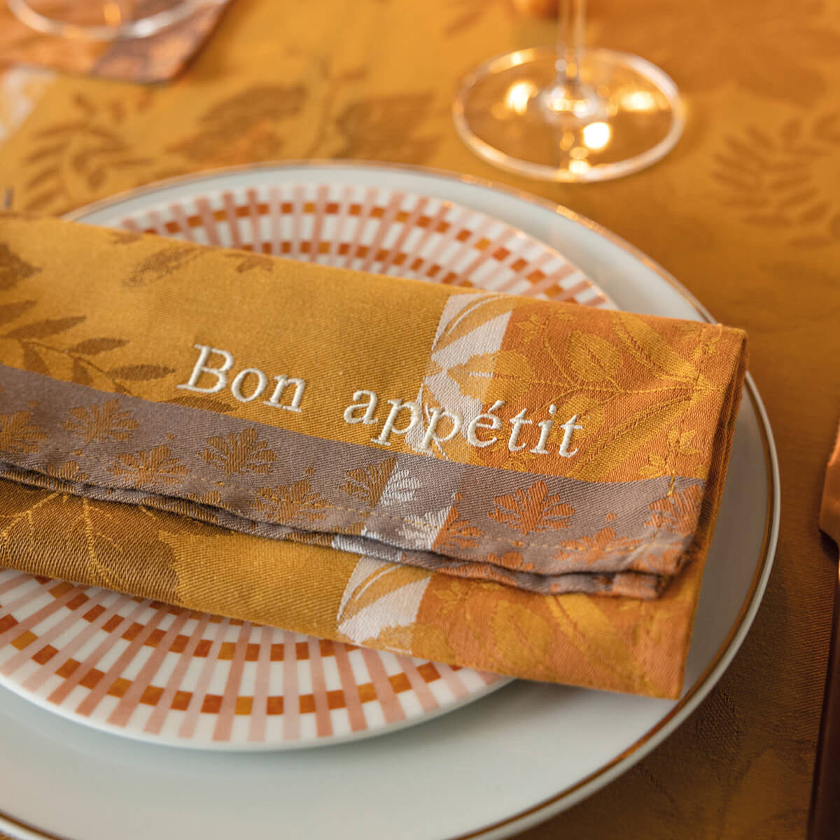 Close-up of folded Orange Manor Lane napkin with "Bon appétit" embroidery on an autumn-toned tablescape.