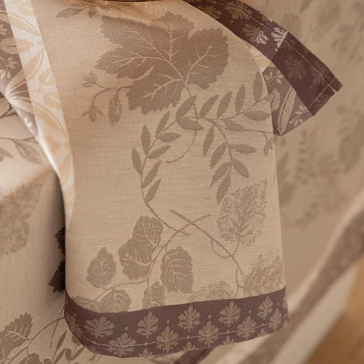 Close-up of Beige Manor Lane dinner napkin showing woven leaf jacquard pattern and contrast border detail.