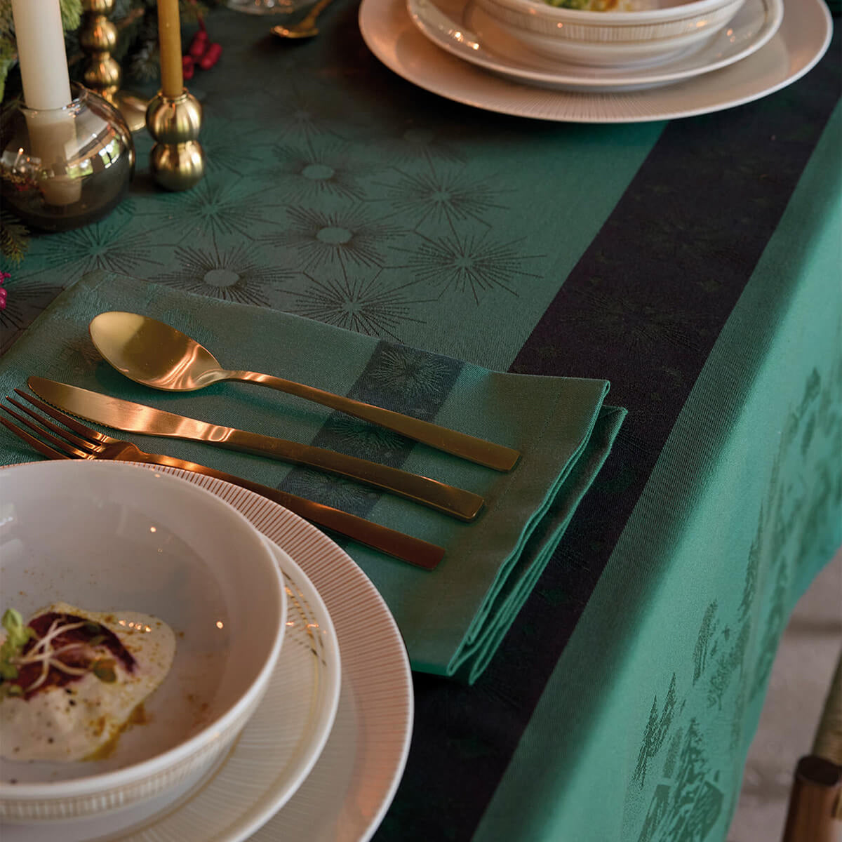 Green jacquard dinner napkin with gold cutlery resting on it, styled on a matching tablecloth.