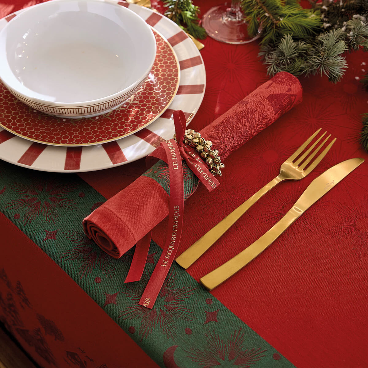 Le Jacquard Français Sous les Étoiles red napkin rolled with ribbon beside gold cutlery on holiday table.