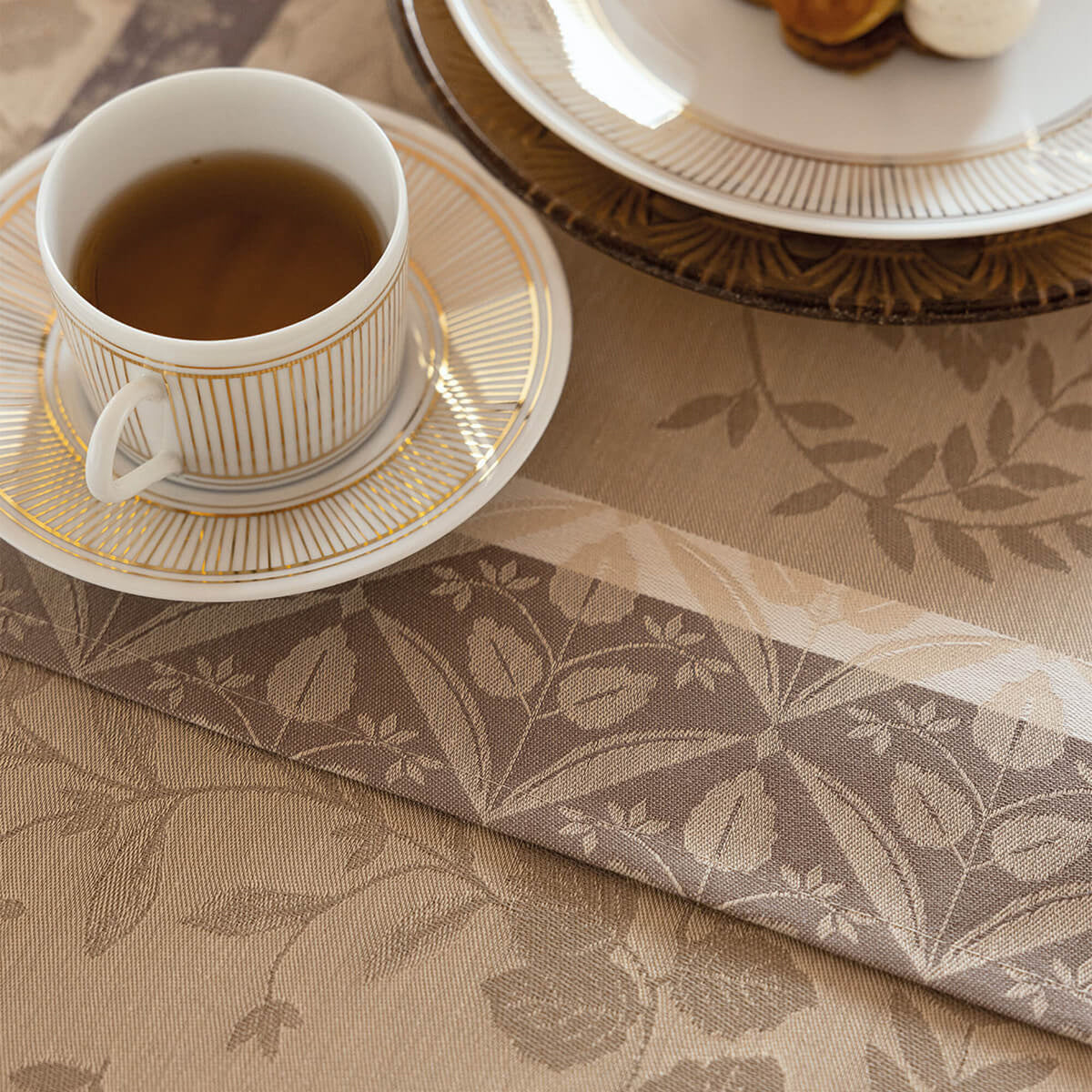 Close-up of Le Jacquard Français Manor Lane placemat in Beige with floral jacquard weave, styled with a gold-striped teacup.