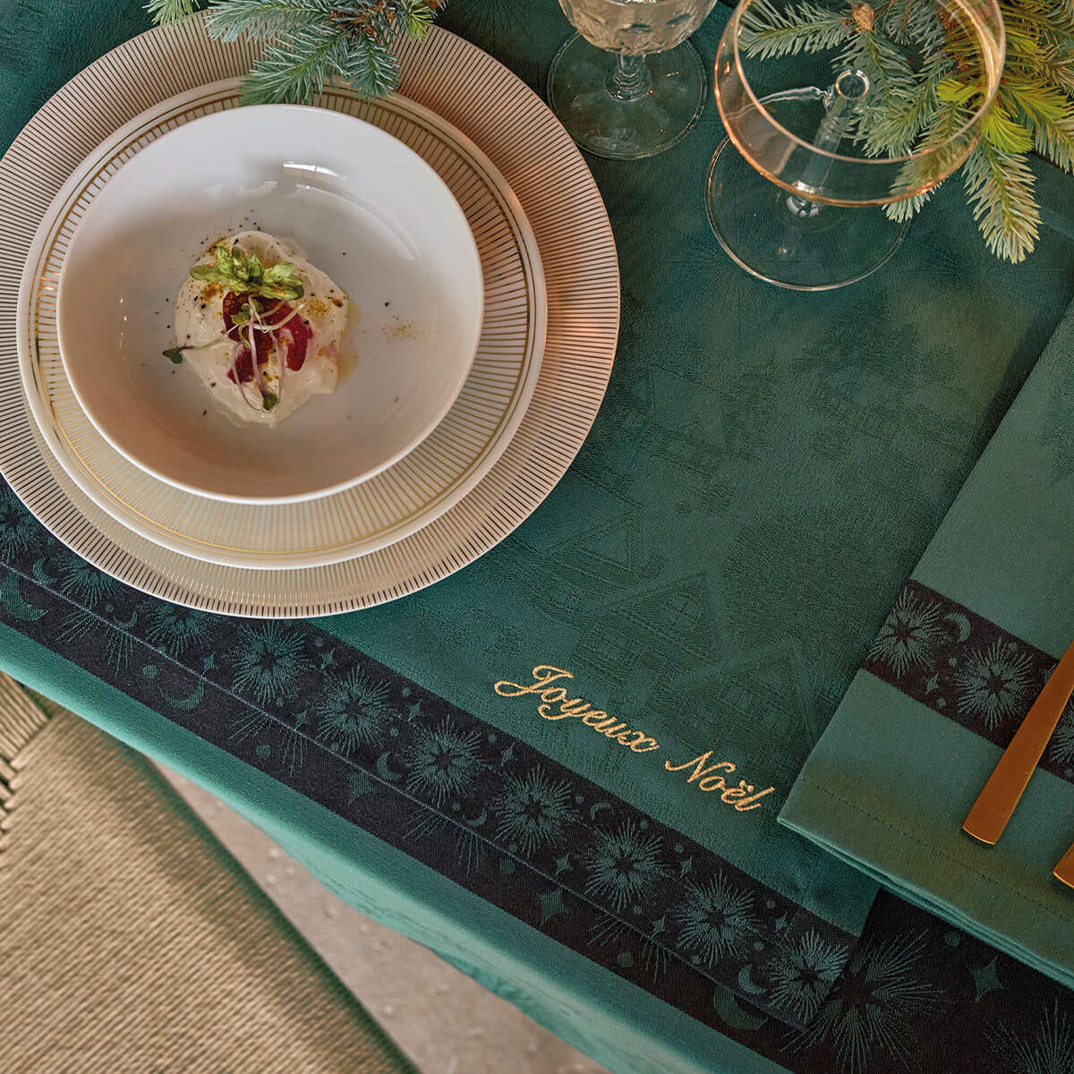 Green placemat with gold "Joyeux Noël" script and dark floral border, styled with fine dining place setting.