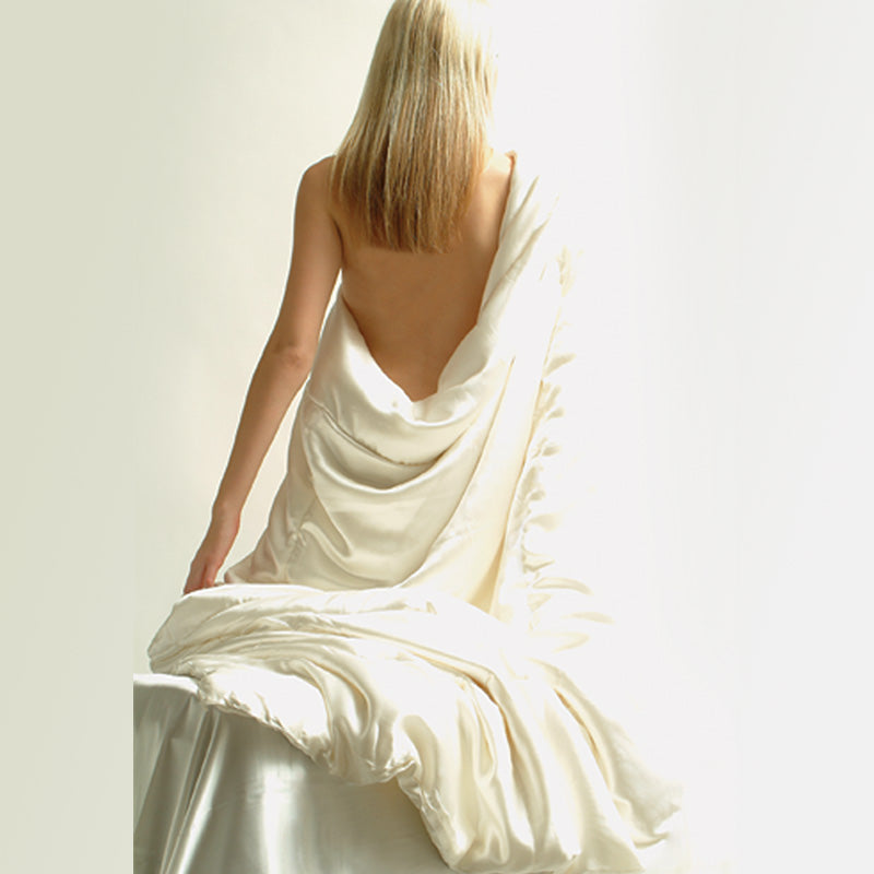 A woman with blonde hair sits with her back exposed, draped in a luxurious Kumi Kookoon Silk Covered Silk-filled Comforter on a white surface against a light background.