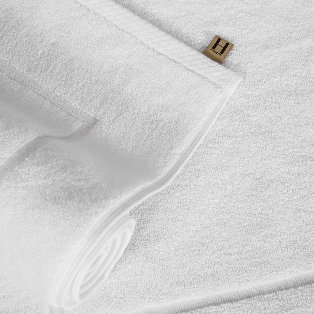Close-up of the Simple Border Bath Mat by Frette, in white, partly rolled, with a small gold and black label with an "H" attached to the edge.