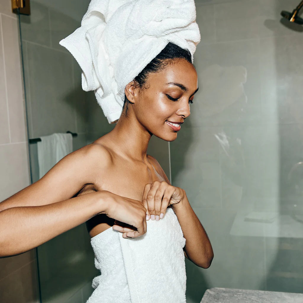 A person stands in a bathroom wrapped in the Simple Border | Bath Sheet Set by Frette, with one towel around their body and another around their head, smiling as they adjust the luxurious Turkish towels.