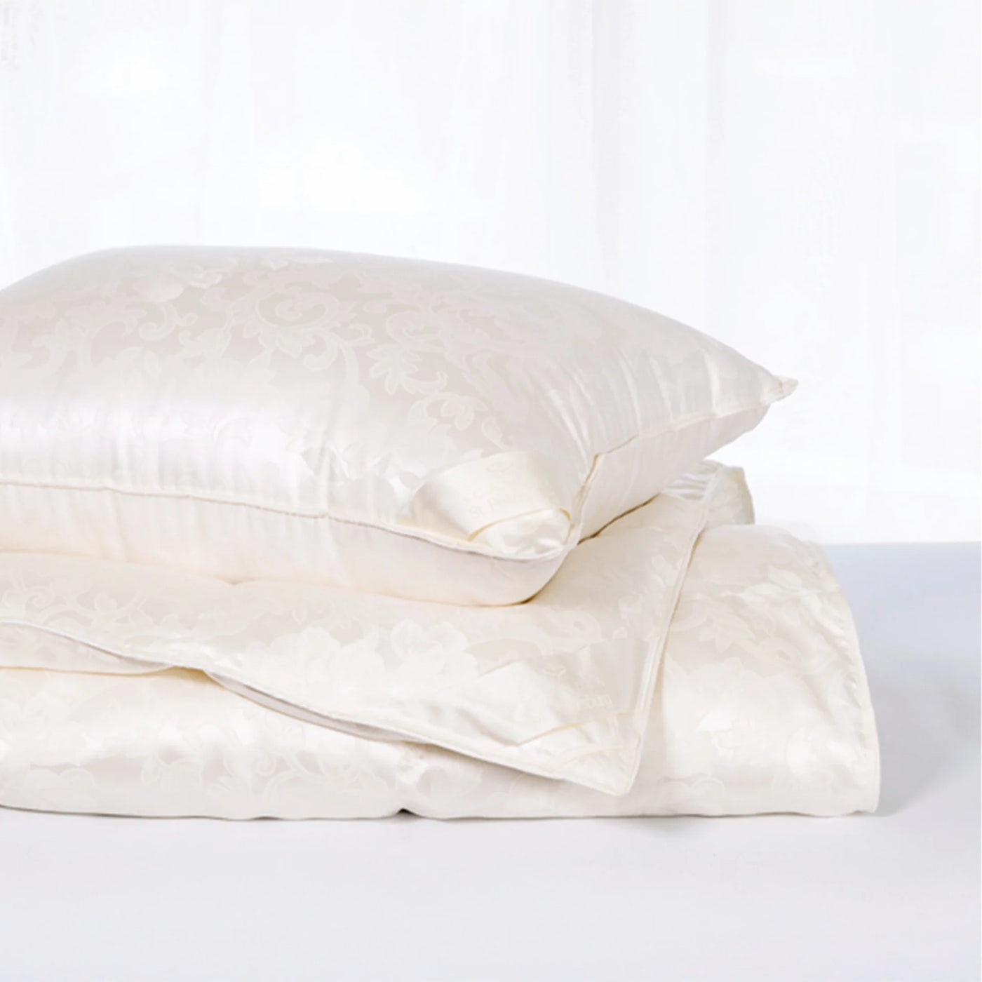 Close-up of ivory silk jacquard pillow stacked on folded bedding, floral woven pattern visible.
