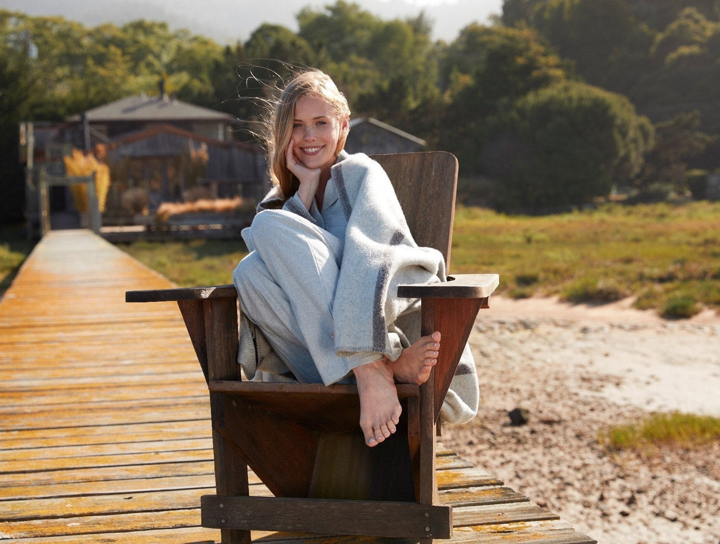 Woman wrapped in Coyuchi Tahoe pale blue wool throw, seated outdoors on wooden dock.