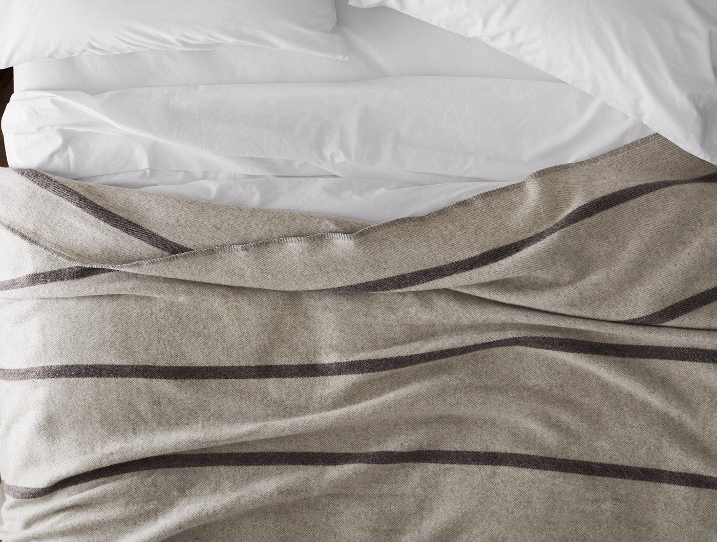 Draped view of oatmeal wool blanket with charcoal stripes over white bedding.