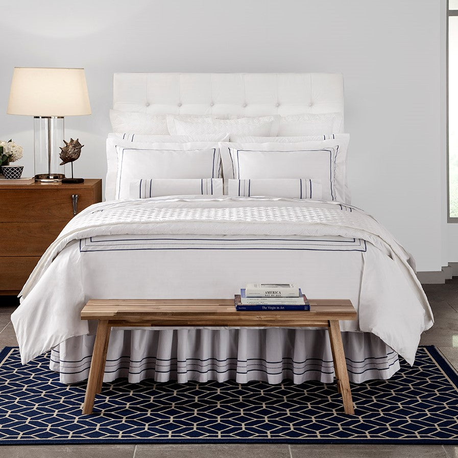 White cotton bedding with navy triple-stripe trim, styled on a bed in a bright modern bedroom.