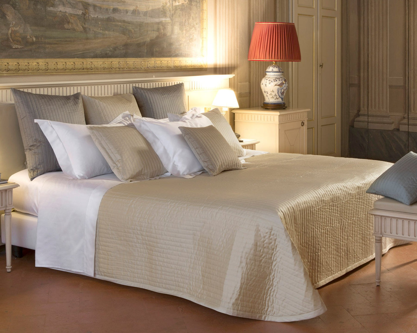 A quilted pillow sham on a bed in a well-lit, classic bedroom decorated with neutral colors and a small red lamp on the nightstand.