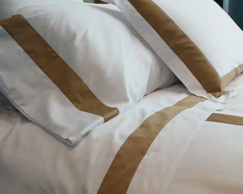 Close-up of white pillowcases with wide gold satin border bands on a made bed.