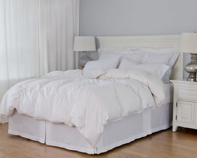A neatly made bed features the St. Geneve Heirloom | Duvet, hypoallergenic pillows, and white bedding, flanked by white nightstands with lamps in a bright room with light walls and curtains.